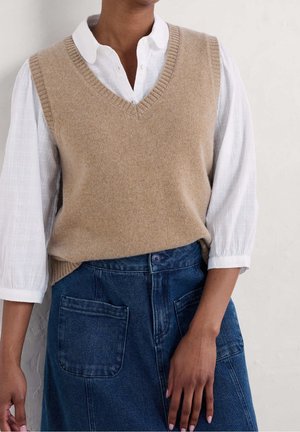 Personne portant un pull sans manches beige en maille par-dessus un chemisier blanc à manches trois-quarts et une jupe en jean bleu avec des poches à l'avant.