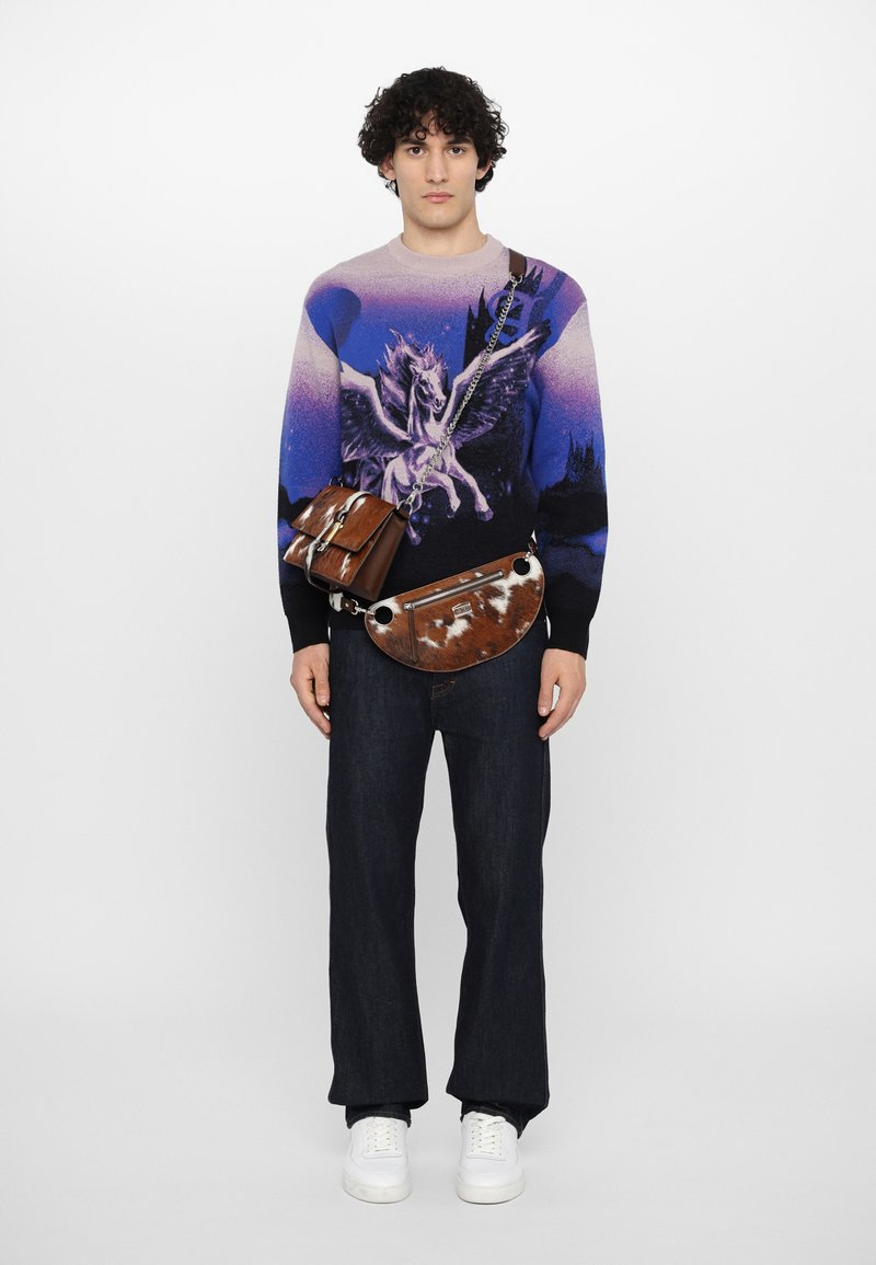 Sweater med en lilla gradient og et unicorn grafik, parret med brune lædertasker og mørke denim jeans. Hvide sneakers fuldender looket.