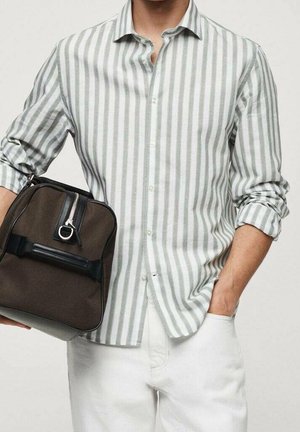 Mężczyzna w jasno szarej i białej koszuli w pionowe paski z podwiniętymi rękawami, białe spodnie, trzymający ciemnobrązową torbę na zamek.