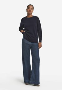 Marineblauer Strickpullover mit geripptem Saum, kombiniert mit weit geschnittenen, dunklen Jeans. Das Model posiert selbstbewusst mit der Hand in der Tasche und trägt goldene Ohrringe.