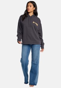 Sudadera con capucha de color gris oscuro, con cordón, bolsillo frontal tipo canguro y logo dorado. Combinada con jeans acampanados de color azul claro.