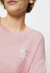 Hellrosa Baumwoll-T-Shirt mit rundem Halsausschnitt und kurzen Ärmeln. Mit weißem Blumenmuster und einem kreisförmigen Akzent auf der Brust.