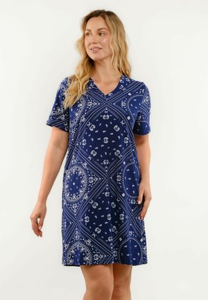 Kobieta z długimi blond włosami, ubrana w niebieską sukienkę z krótkim rękawem, z białym wzorem paisley i geometrycznymi, stojąca na jednolitym tle.