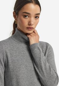 Grauer Rollkragenpullover aus weichem Strick, mit einem gerippten Kragen und Bündchen. Nahaufnahme zeigt die glatte Textur und klare Linien. Akzent mit goldenen Ohrringen.