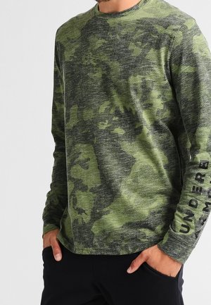 Chemise à manches longues camouflage verte en matériau texturé, avec un col rond et un texte imprimé noir le long de la manche gauche.