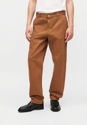 Muž oblečený v voľných hnedých nohaviciach a čiernych kožených topánkach, stojaci proti jednoduchému bielemu pozadiu.