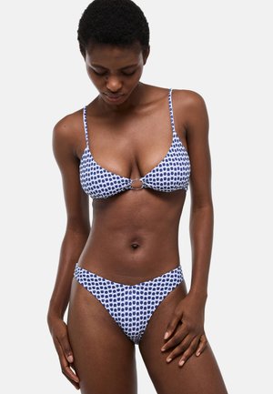 Femme portant un bikini bleu et blanc à pois, regardant vers le bas, debout devant un fond clair uni.