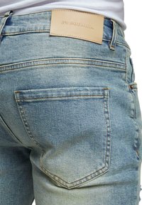 Vaqueros de denim azul claro con un bolsillo trasero con costuras visibles y una etiqueta de marca de cuero beige en la cinturilla.