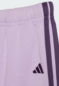 Un pantalon de survêtement lavande avec une taille ornée de trois rayures violettes sur le côté et un petit logo Adidas près du bord de la poche.