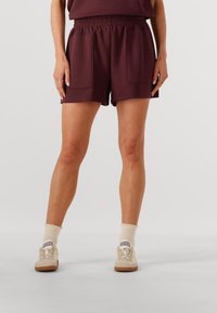 Pantalones cortos burdeos con cinturilla elástica, dos bolsillos frontales y un corte relajado. Llevados con zapatos de color claro y calcetines tobilleros.