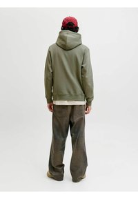 Felpa con cappuccio verde oliva con vestibilità rilassata, abbinata a pantaloni di jeans grigi larghi e un cappellino rosso, che mostra un design casual e texture morbide.