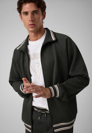 Jeune homme portant une veste noire zippée avec des poignets et un col rayés blancs sur un t-shirt blanc à motif graphique, debout devant un fond gris.