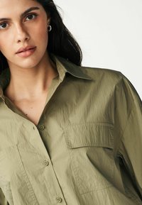 Khaki košile s knoflíky a jemnou strukturou, která má strukturovaný límec a dvě náprsní kapsy. Materiál má mírně pomačkaný vzhled.