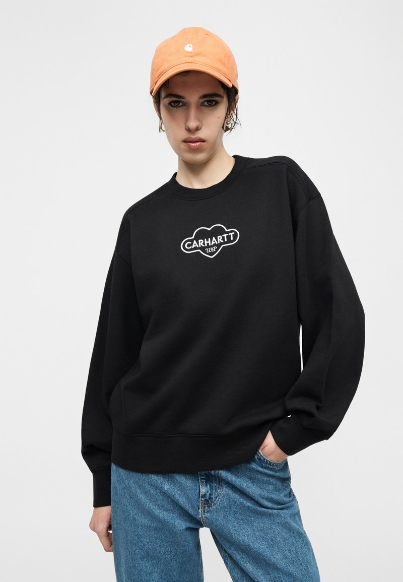 Persona con cappellino arancione, felpa Carhartt nera con logo e jeans blu, in piedi con una mano in tasca contro uno sfondo semplice.