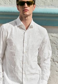 Witte overhemd met textuur en bloemenborduursel, lange mouwen en een klassieke kraag. Model draagt groene zonnebril.