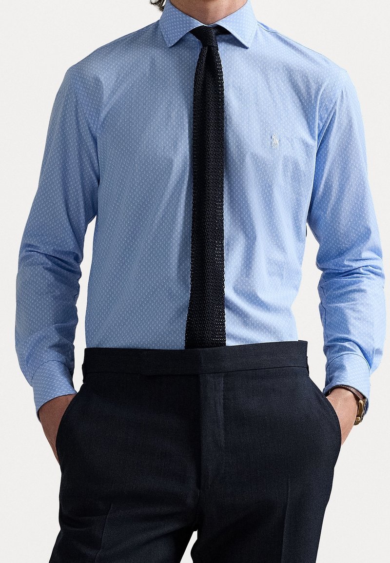 Mies, joka on pukeutunut vaaleansiniseen kuvioituun kauluspaitaan, mustaan teksturoituun solkeen, mustiin housuihin, kädet taskuissa ja ruskeaan nahkarannekelloon.