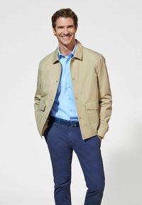 Beżowa, lekka kurtka z zatrzaskami, z dwiema przednimi kieszeniami, noszona na jasnoniebieską koszulę, połączona z granatowymi spodniami i czarnym paskiem.