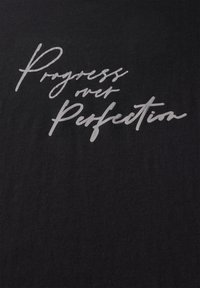 Musta t-paita, jossa on valkoinen käsinkirjoitustyylinen tekstigraafi, jossa lukee "Eteneminen yli täydellisyyden" edessä. Pehmeä kangas.