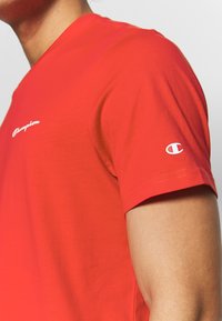 Rotes Baumwoll-T-Shirt mit Rundhalsausschnitt, kurzärmlig und mit einem weißen "Champion"-Logo auf der Brust und dem Ärmel. Glatte Textur.