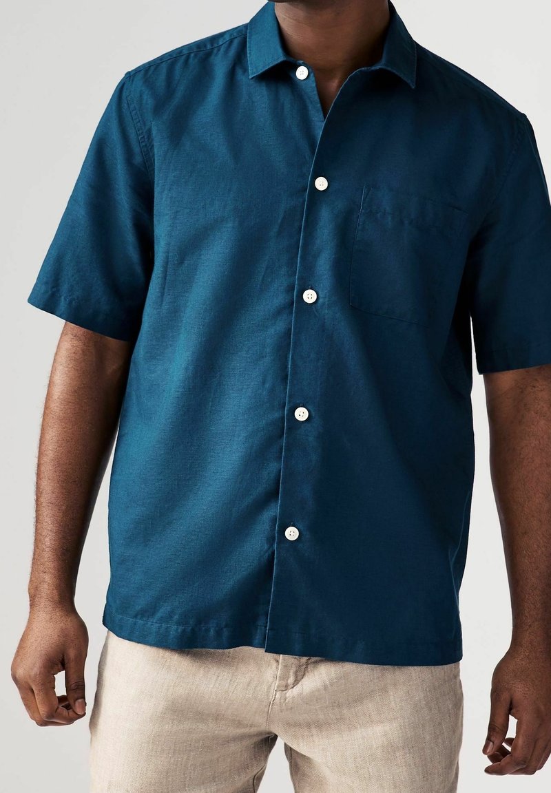 Homme portant une chemise à manches courtes boutonnée bleu foncé et un pantalon beige clair, debout devant un fond uni.