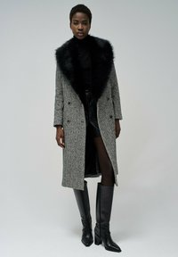 Cappotto grigio a motivo houndstooth con colletto in pelliccia sintetica nera, design doppiopetto, abbinato a un top nero, gonna di pelle e stivali al ginocchio.