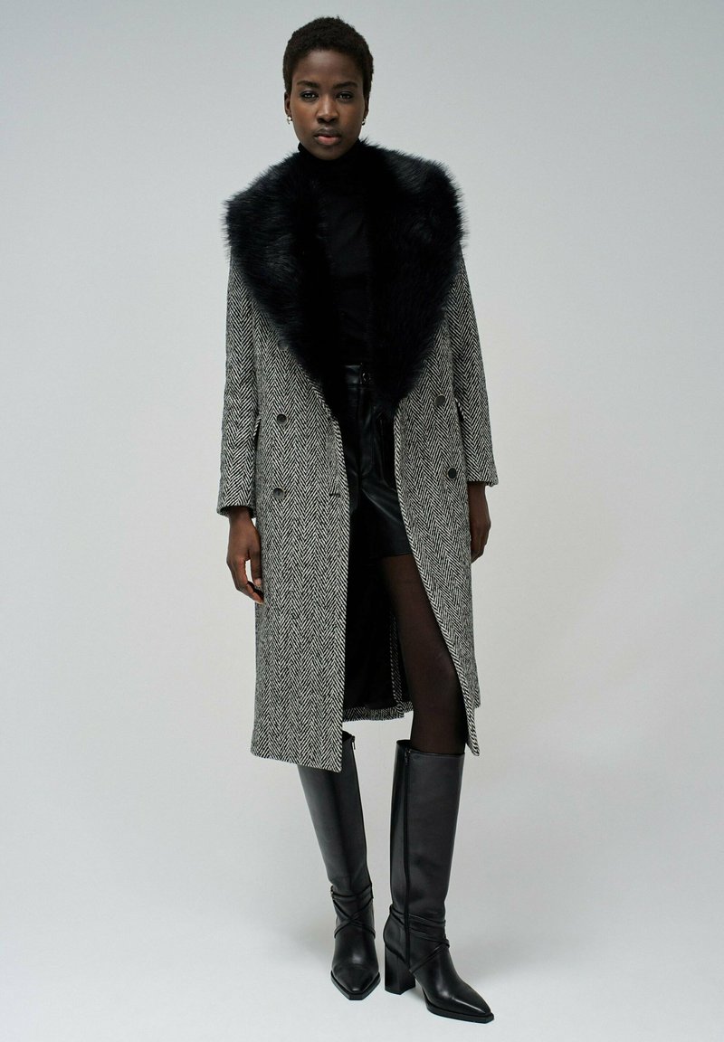 Cappotto grigio a motivo houndstooth con colletto in pelliccia sintetica nera, design doppiopetto, abbinato a un top nero, gonna di pelle e stivali al ginocchio.