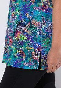 Hånd hvilende ved siden af en farverig tunika med blomsterprint og sidelommer, kombineret med sorte bukser.