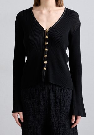 Kobieta ubrana w czarny, prążkowany, długorękawowy cardigan z siedmioma brązowymi guzikami z przodu, nosząca teksturowane czarne spodnie.