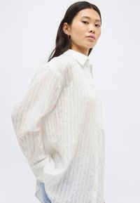 Mujer joven con cabello largo y oscuro lleva una camisa blanca semitransparente y suelta con botones y rayas verticales, posando con las manos en los bolsillos sobre un fondo blanco.