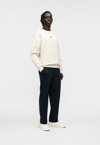 Sweat-shirt crème avec logo noir, associé à un pantalon bleu foncé. Le modèle porte des baskets blanches. Tissu lisse avec une coupe décontractée et un design simple.