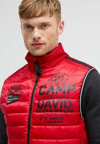 Rote gepolsterte Jacke mit hohem Kragen, ausgestattet mit schwarzen Textdrucken und einem Reißverschluss. Das Material wirkt glatt, mit einem kontrastierenden Design der schwarzen Ärmel.