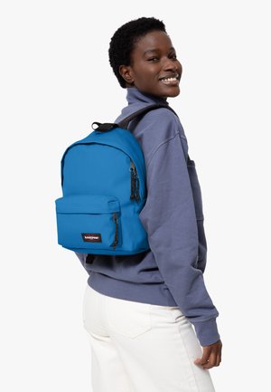 Mochila azul con una tela suave y duradera, que cuenta con un bolsillo frontal, cremalleras negras y un parche con el logo. Lleva puesta a la espalda de una persona.