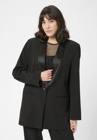 Fekete blazer selyem reverrel, strukturált kialak�ítással, elöl gombos záródással, két zsebbel, fekete, áttetsző pöttyös blúz felett viselve.