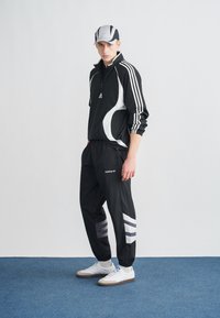 Μαύρη αθλητική φόρμα με λευκές λεπτομέρειες και τρεις ρίγες. Ελαφρύ ύφασμα, φερμουάρ στον γιακά και παντελόνι με στενή γραμμή και ρίγες. Λευκά αθλητικά παπούτσια.