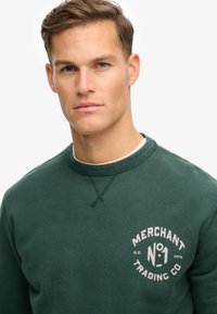 Sweat-shirt vert à col rond, avec un tissu texturé et un logo blanc sur la poitrine indiquant "MERCHANT TRADING CO No. 1".