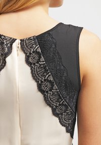 Černá krajková akcentace na rameni spojuje krémovou látku se zipovým uzávěrem vzadu. Měkká textura a přiléhavý design zvýrazňují oděv.