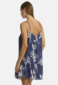 Una mujer con cabello largo lleva un vestido sin mangas de color marino con un patrón floral. Se muestra de espaldas, destacando el corte y diseño del vestido.