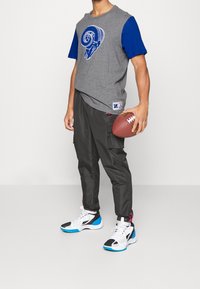 Camiseta gris con mangas azules y un diseño de carnero, pantalones cargo negros y zapatillas blancas con acentos en azul y negro. Sosteniendo un balón de fútbol.