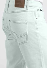 Jean vert clair en denim, présentant des coutures contrastantes, une étiquette de marque en cuir marron, et deux poches arrière aux bords arrondis.