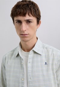 Jasno-koszula w kratę z niebieskimi liniami, kołnierzykiem na guziki i małym haftowanym logo czaszki na lewej stronie klatki piersiowej.