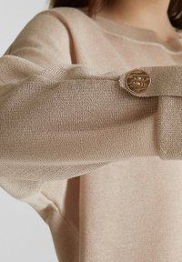 Beżowy sweter dziany z szerokim mankietem, zapinanym na teksturowany, okrągły złoty guzik.