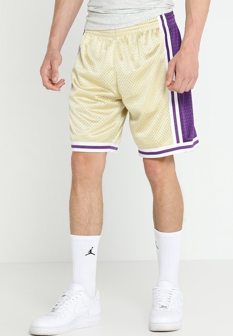 Homme portant un short de basket beige avec des rayures violettes et blanches, des chaussettes blanches montantes avec un logo noir, et des baskets blanches.