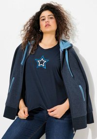 Námořnická modrá mikina na zip s světle modrými akcenty, spárovaná s námořnickým tričkem s bílým a světle modrým hvězdným designem. Tmavé džíny.