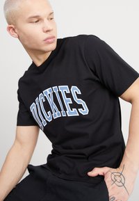 Černé bavlněné tričko s výrazným bílým a světle modrým nápisem "DICKIES", krátkými rukávy a volným střihem.