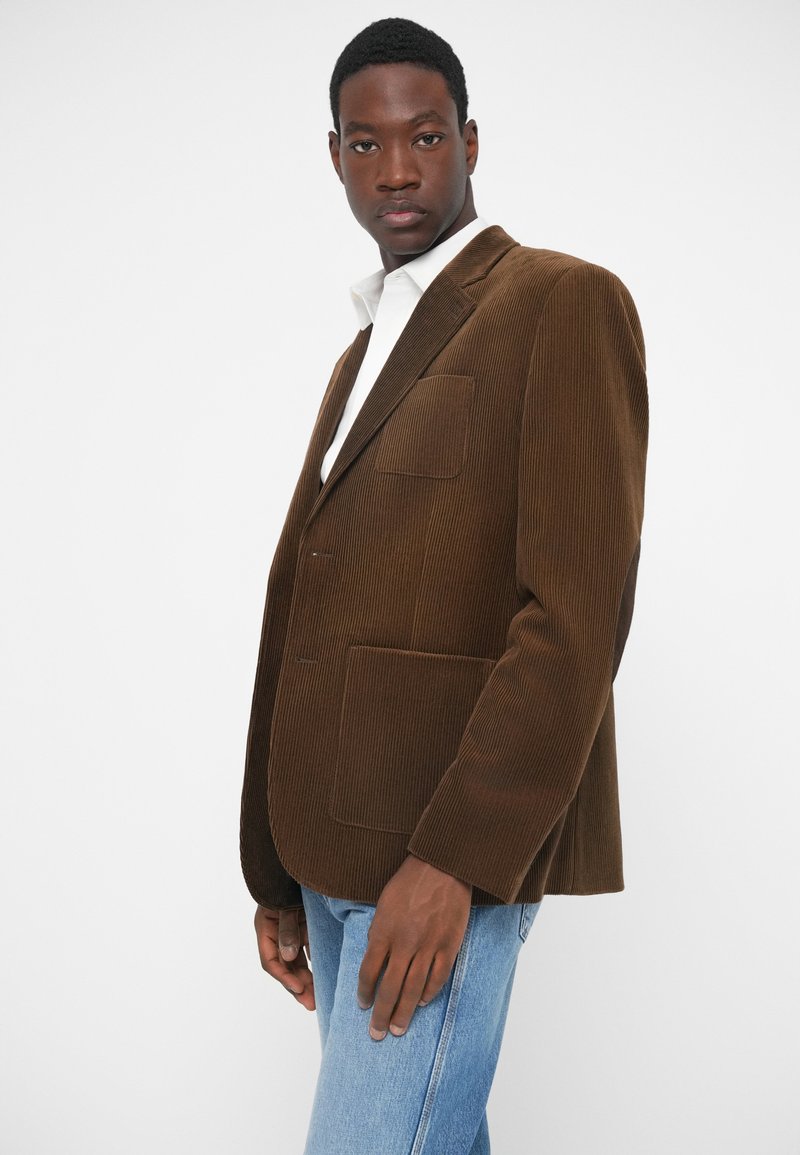 Brun kordflädsblazer med en strukturerad design, som har en klädd krage och två framfickor. Styld över en vit skjorta och blå jeans.