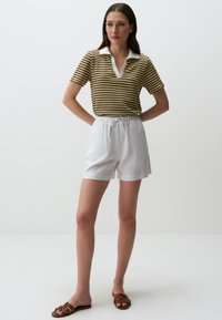 Polo a righe con maniche corte in verde, giallo e marrone, con colletto bianco, abbinato a pantaloni corti bianchi con cordino e sandali marroni.