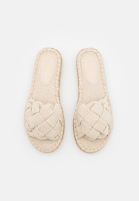 Sandales à glisser tissées de couleur beige avec une sangle tressée, semelle plate et surface en tissu texturé. Présente un bord cousu autour de la base.
