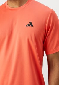 Cămașă sport coral, realizată dintr-un material texturat și respirabil, având mâneci scurte și un logo negru accentuând partea stângă sus.