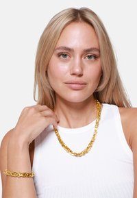 Goldfarbene Kettenanhänger- und Armband-Set mit miteinander verbundenen Gliedern, das ein glattes, glänzendes Finish vor einem weißen Oberteil zeigt.