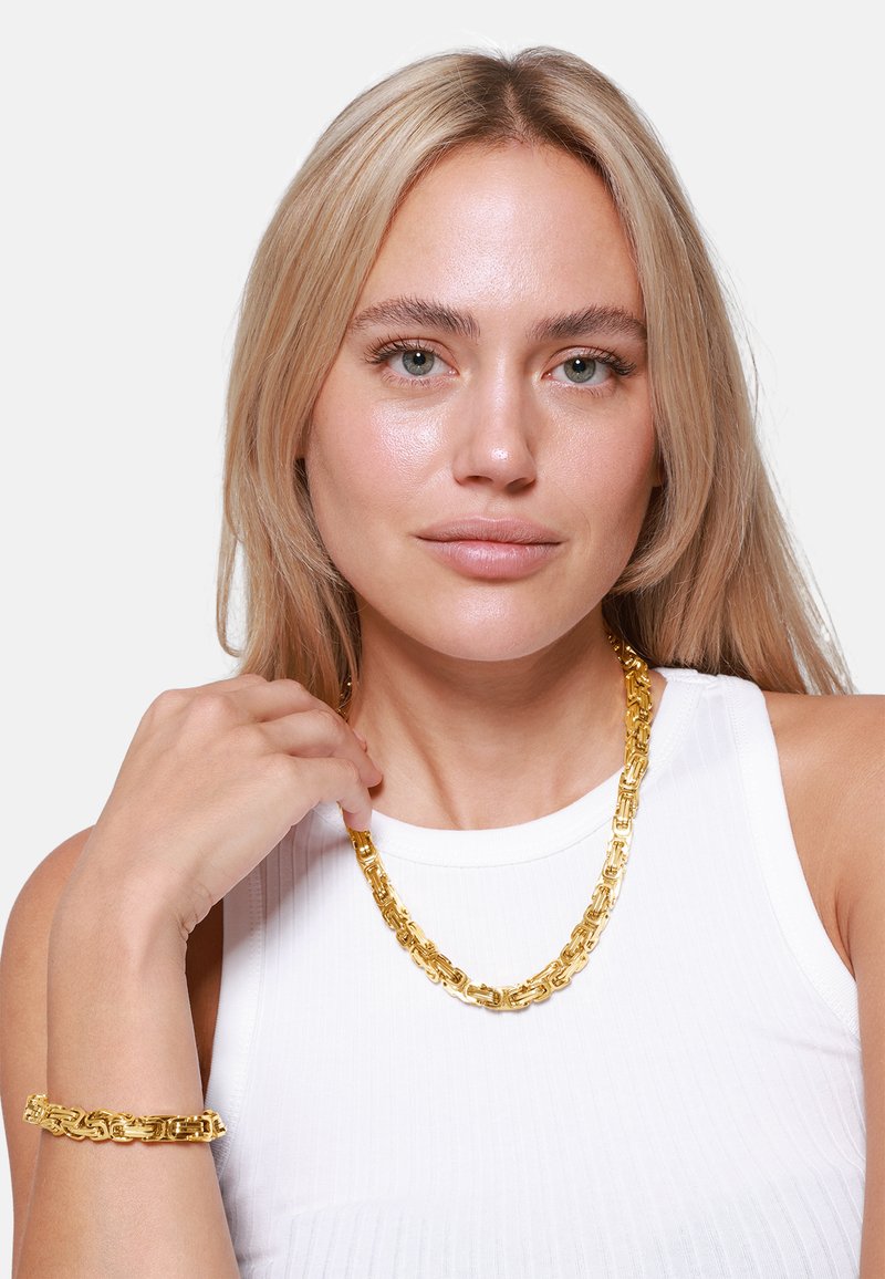 Goldfarbene Kettenanhänger- und Armband-Set mit miteinander verbundenen Gliedern, das ein glattes, glänzendes Finish vor einem weißen Oberteil zeigt.